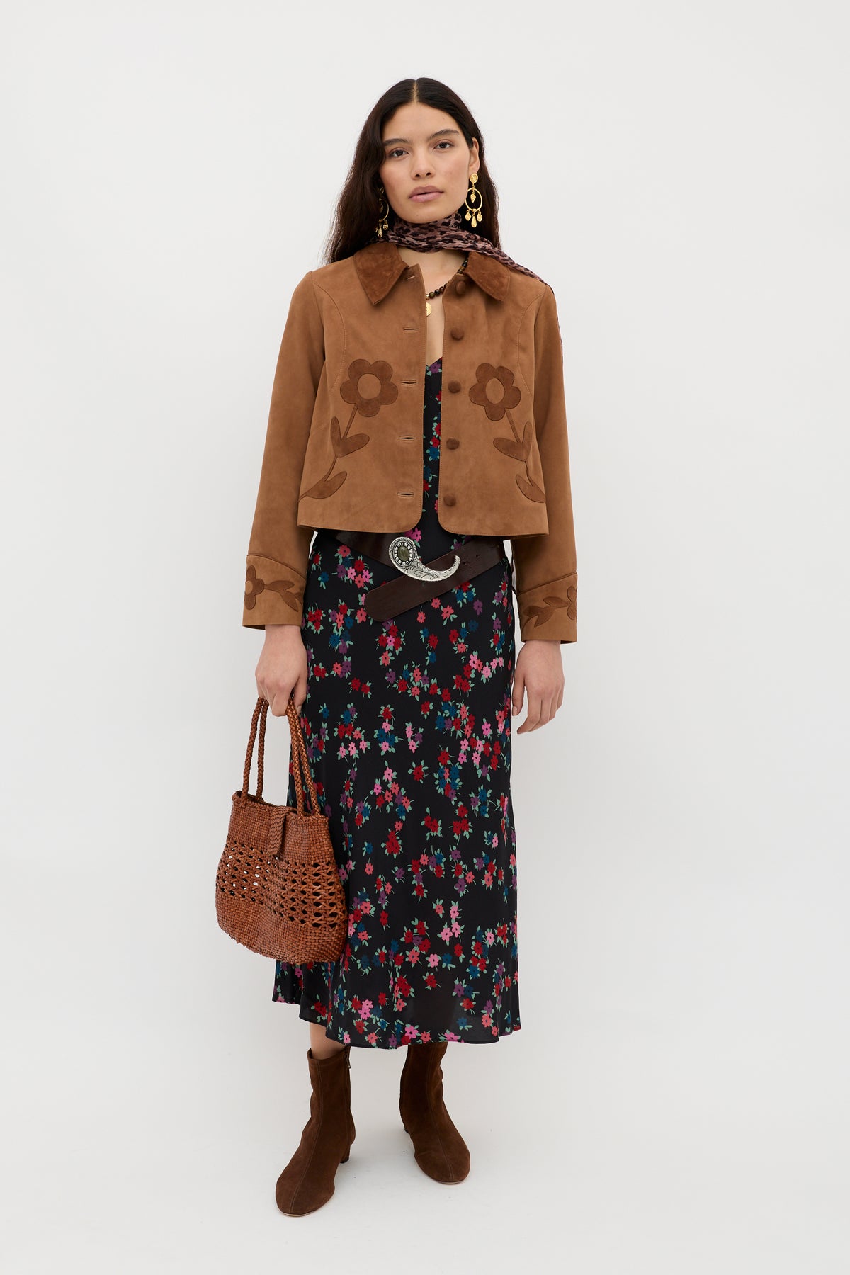 A woman stands against a white background wearing the Mavis - City Floral Black dress, styled with a brown suede jacket, brown boots, a printed scarf, large earrings, a wide belt, and holding a woven brown handbag.