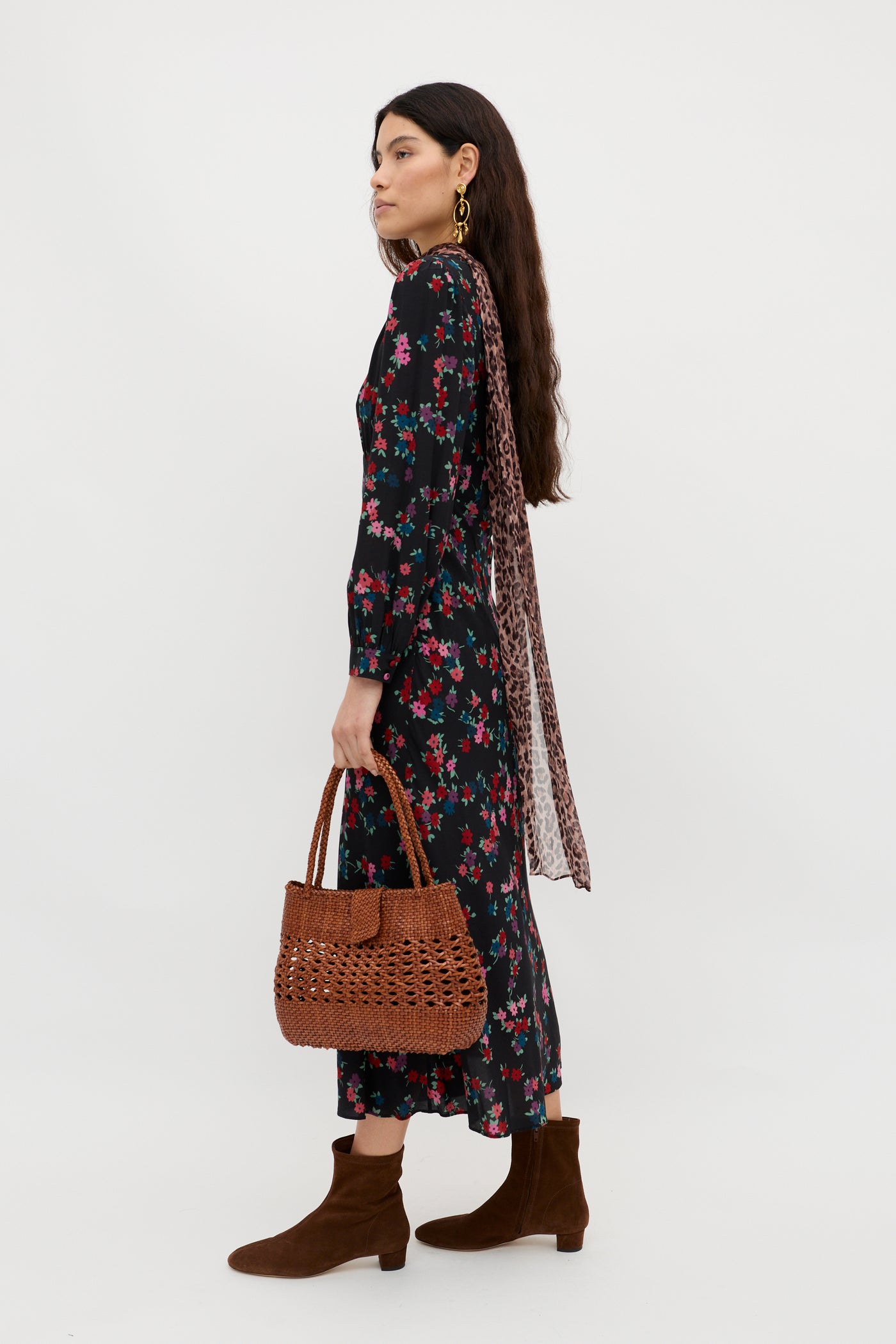 Woman standing sideways against a plain background wears the Mavis - City Floral Black long-sleeve dress—an ideal occasion dress—with brown ankle boots, a brown woven handbag, gold earrings, and a leopard print scarf.