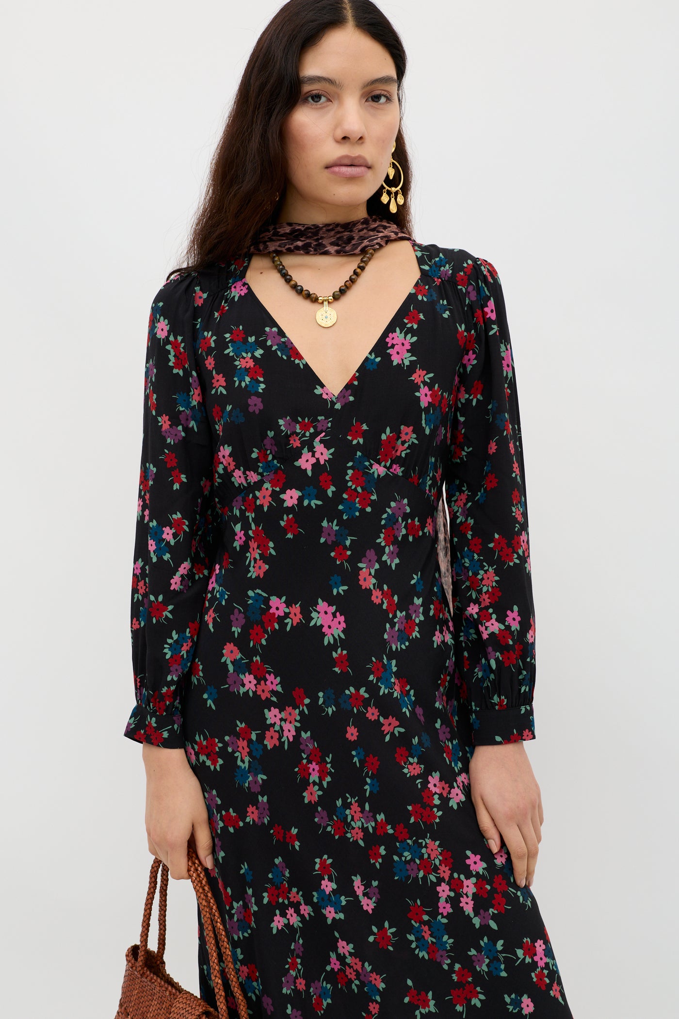 A woman with long dark hair wears the Mavis - City Floral Black dress, gold earrings, a gold necklace, and carries a brown woven bag. She stands against a plain white background.