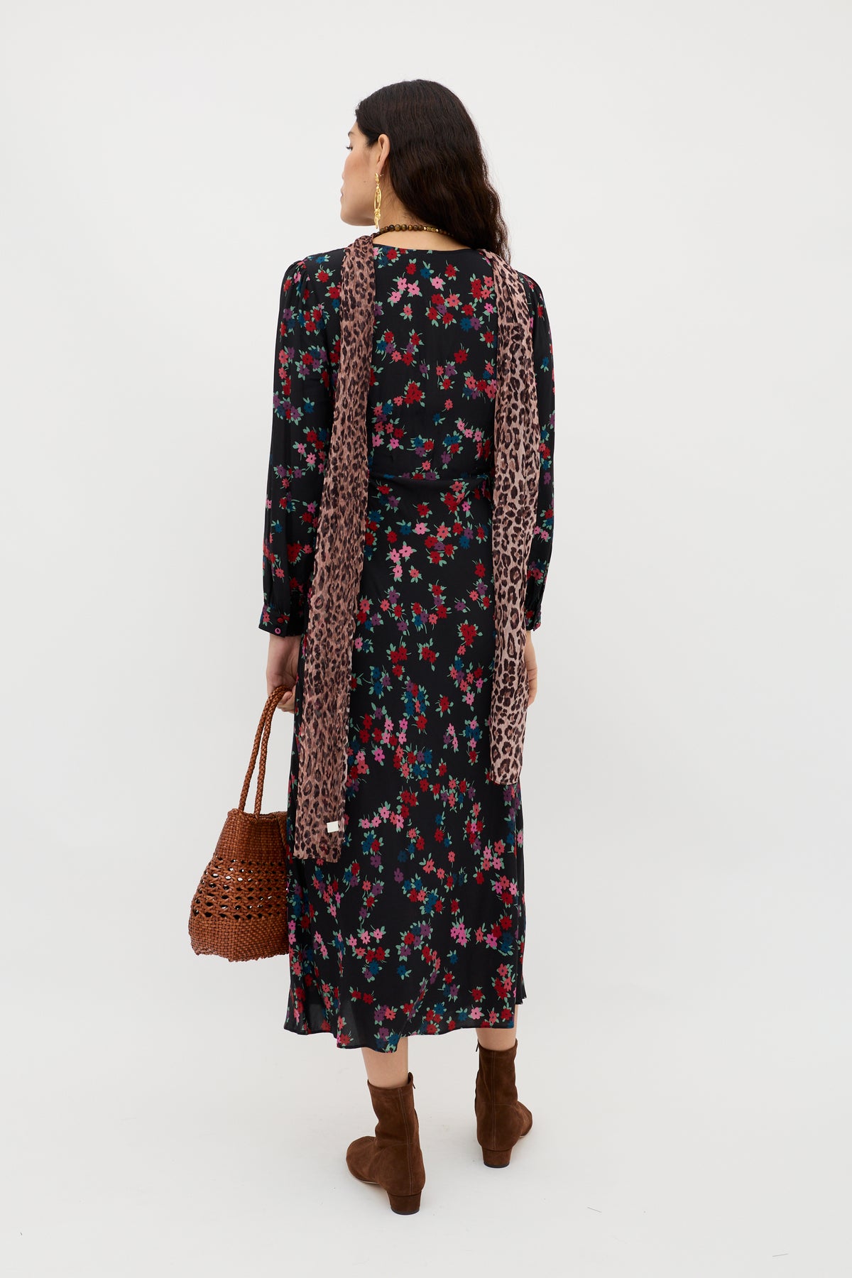 A woman with long dark hair and gold earrings stands facing away against a white background, wearing the Mavis - City Floral Black silk Crepe de Chine dress, brown suede boots, a leopard-print scarf, and carrying a woven brown handbag.
