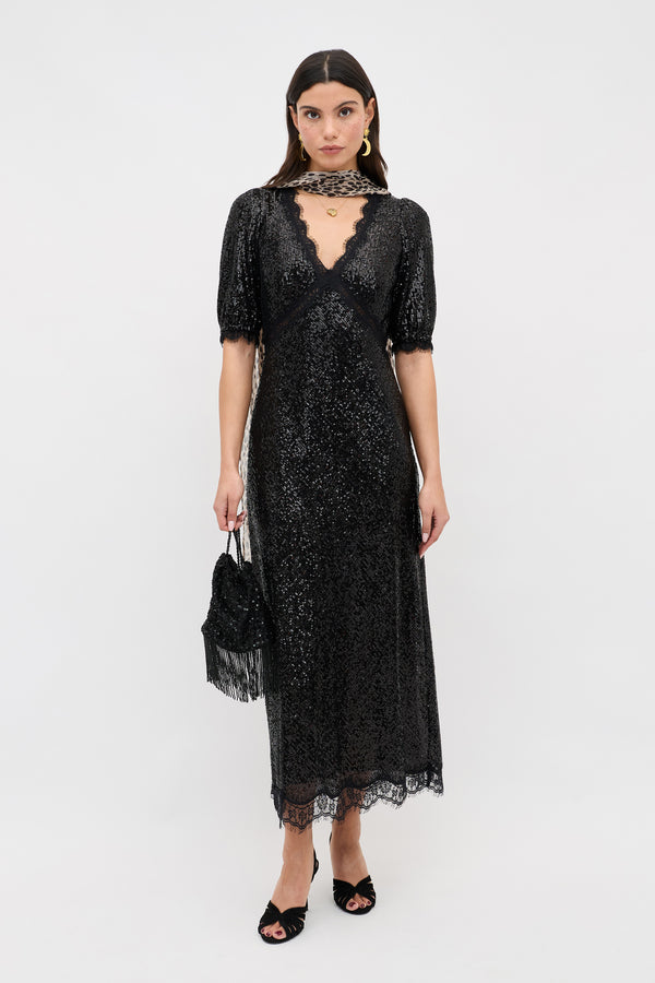 A woman wearing the Verena - Embellishment Black dress, a sparkly black lace midi with short sleeves and a deep V neckline, holds a small black handbag and wears black open-toe heels while standing against a plain white background.