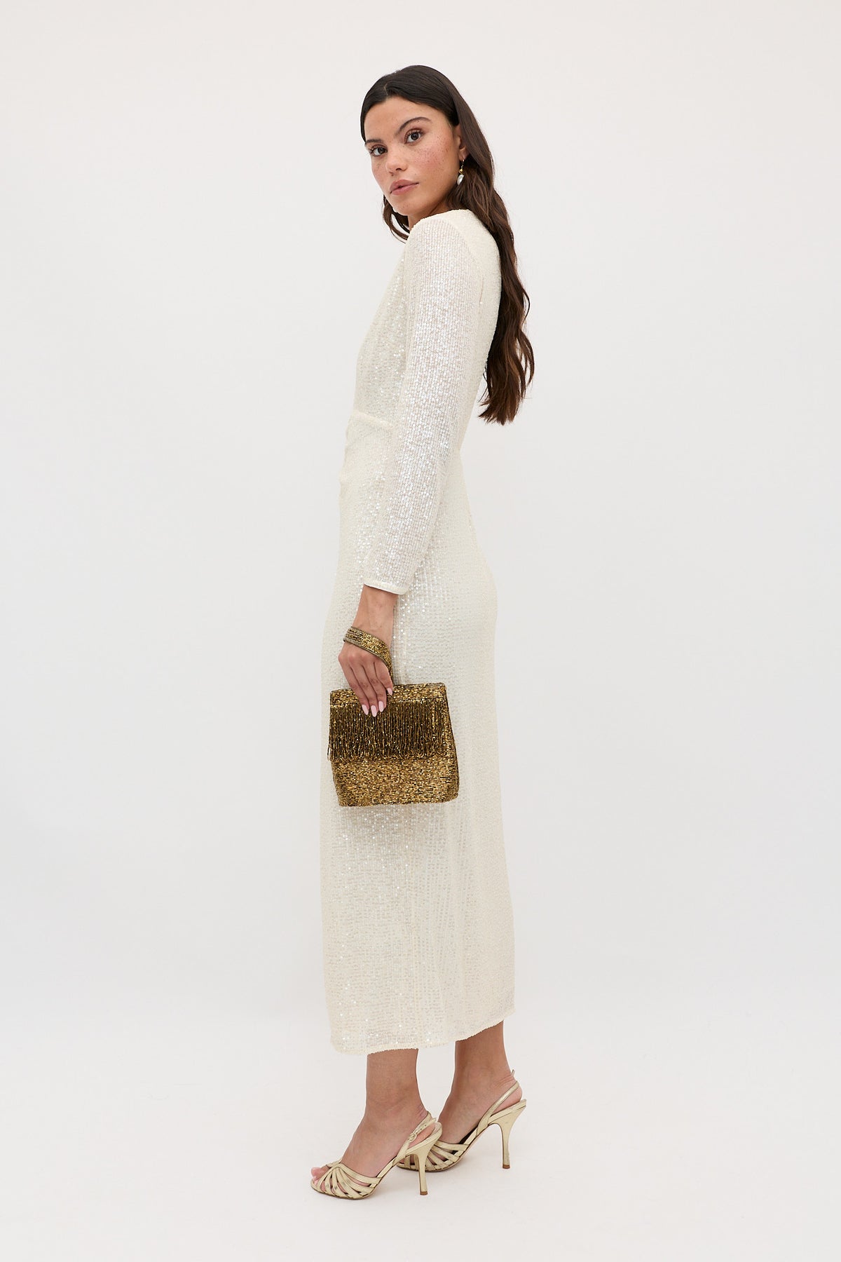 A woman stands sideways in the Lorenzo - Pearl long-sleeved, shimmery ivory dress, holding a gold beaded clutch. She wears heeled sandals, has long wavy brown hair, and poses against a plain white background.