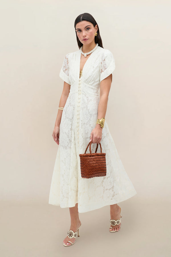 A woman in the Novara - Ivory vintage dress with floral lace and curved sleeves stands against a beige background, holding a woven brown handbag, wearing white heeled sandals and gold jewelry, with a neutral expression.