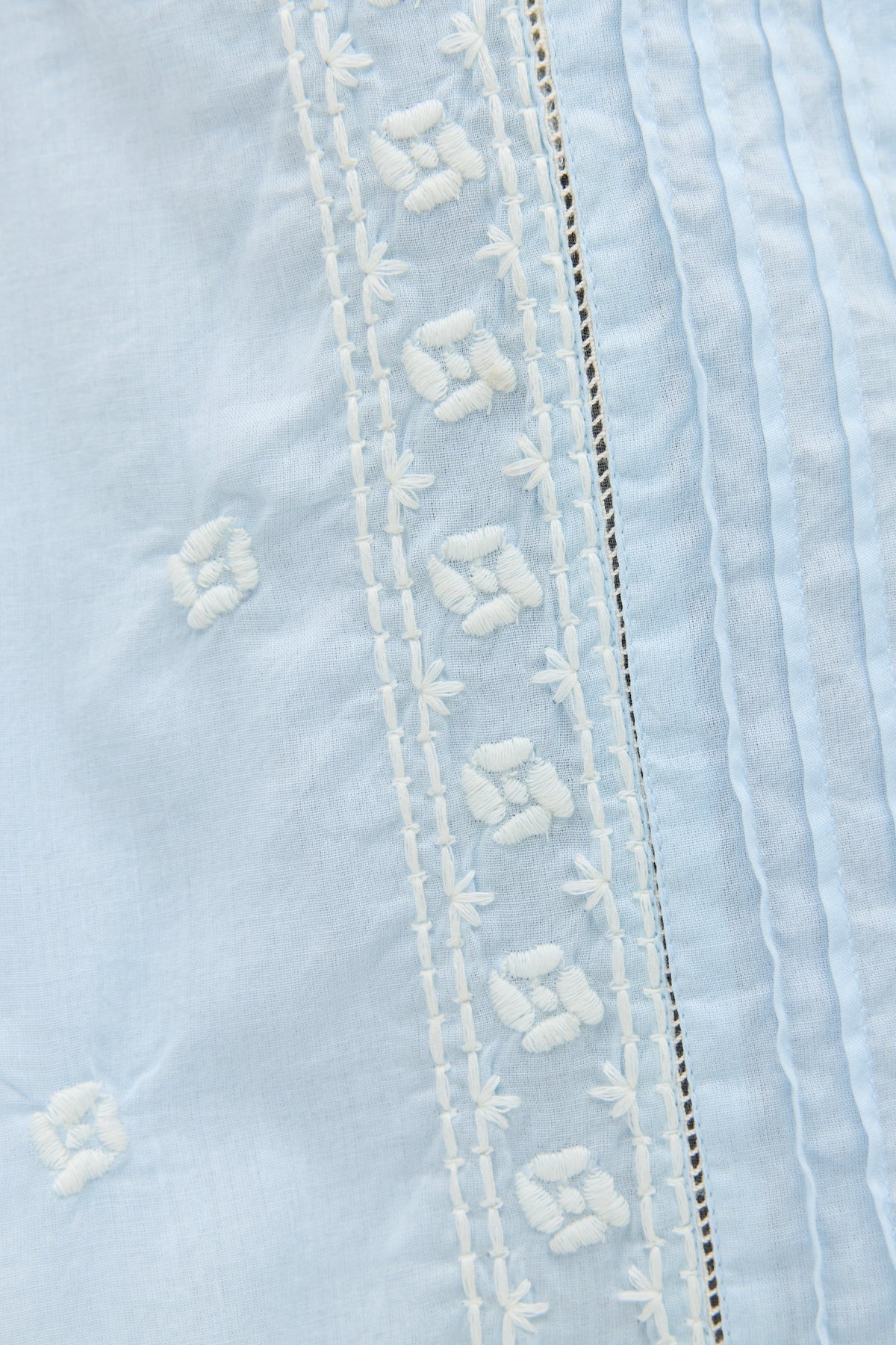 Close-up of the Edina - Mira Embroidery Powder Blue blouse in light cotton-voile, featuring white floral embroidery and vertical pleats.