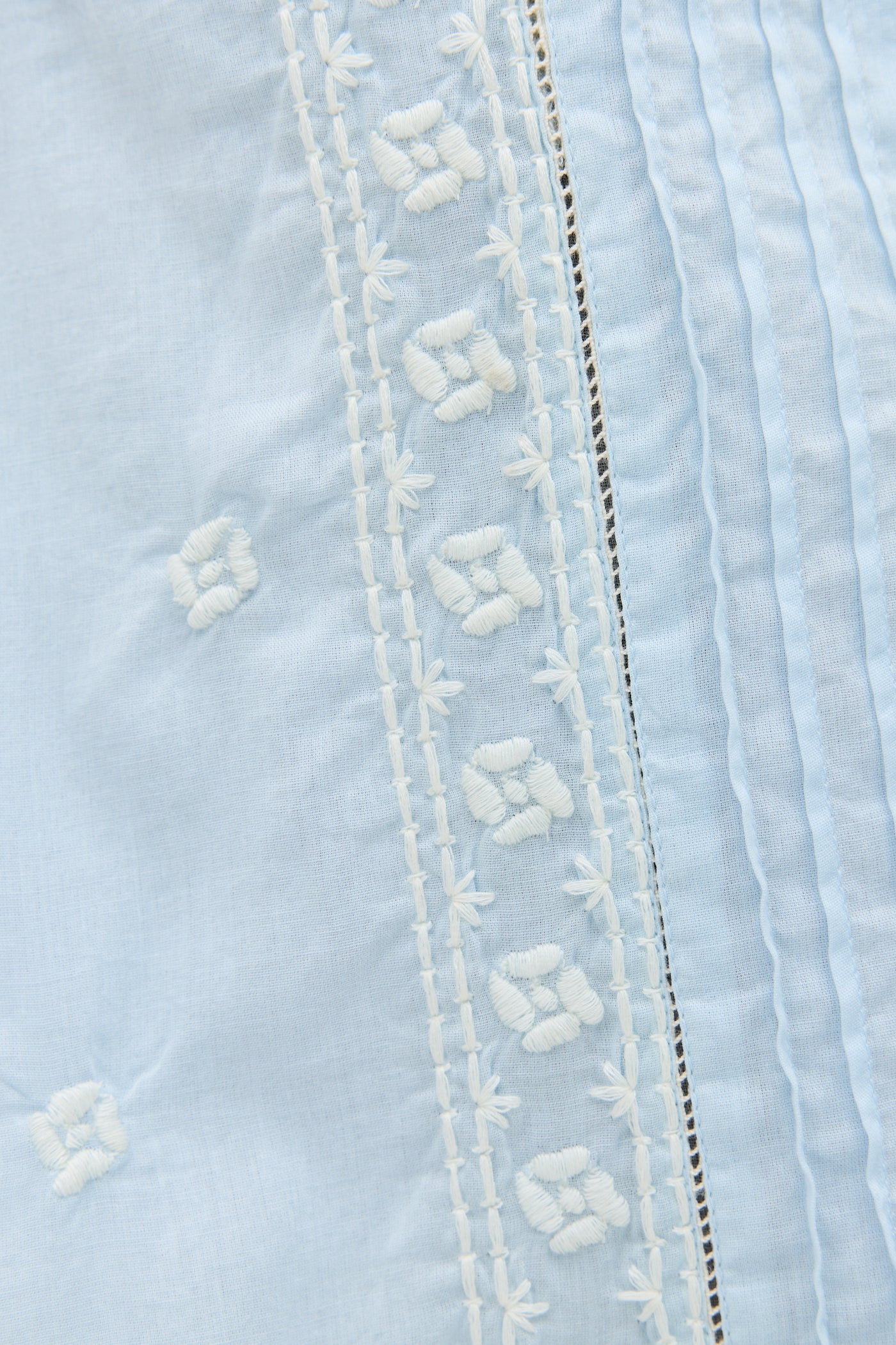 Close-up of the Edina - Mira Embroidery Powder Blue blouse in light cotton-voile, featuring white floral embroidery and vertical pleats.