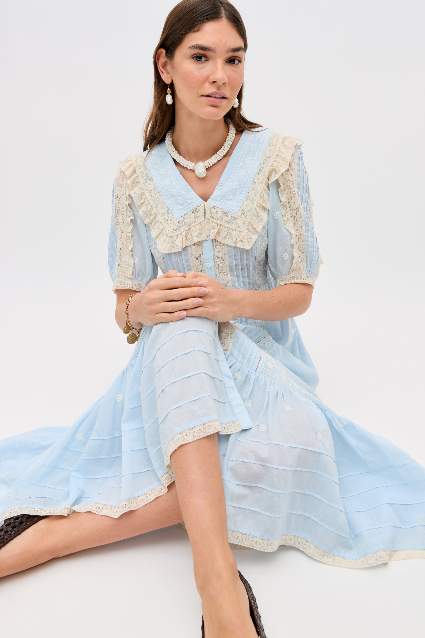 A woman with long brown hair and pearl jewelry sits on the floor in the Edina - Mira Embroidery Powder Blue blouse, featuring lace trim and a large collar, vintage-inspired cotton-voile, against a plain light background.