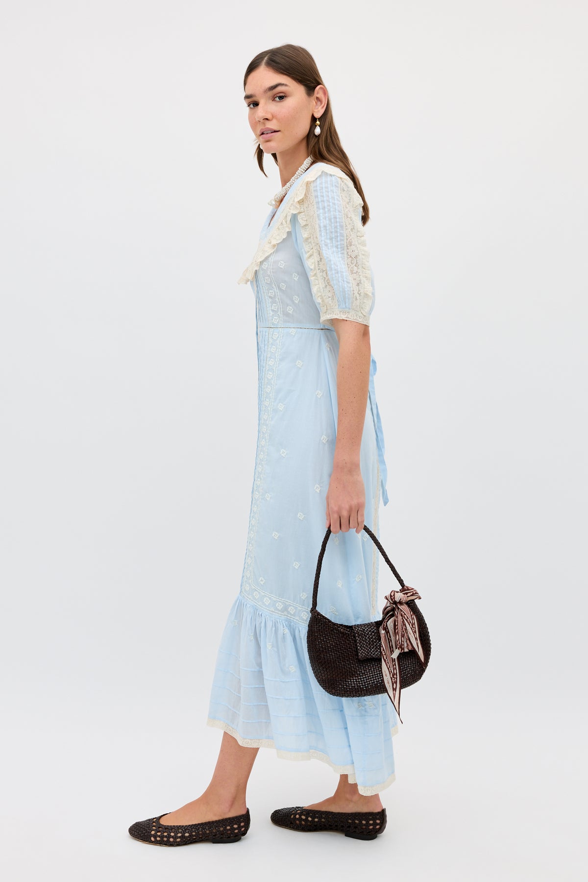 A woman stands sideways in the Edina - Mira Embroidery Powder Blue midi dress with lace details and puff sleeves, holding a dark woven handbag with a patterned scarf. She wears black woven flats and has long, straight hair.