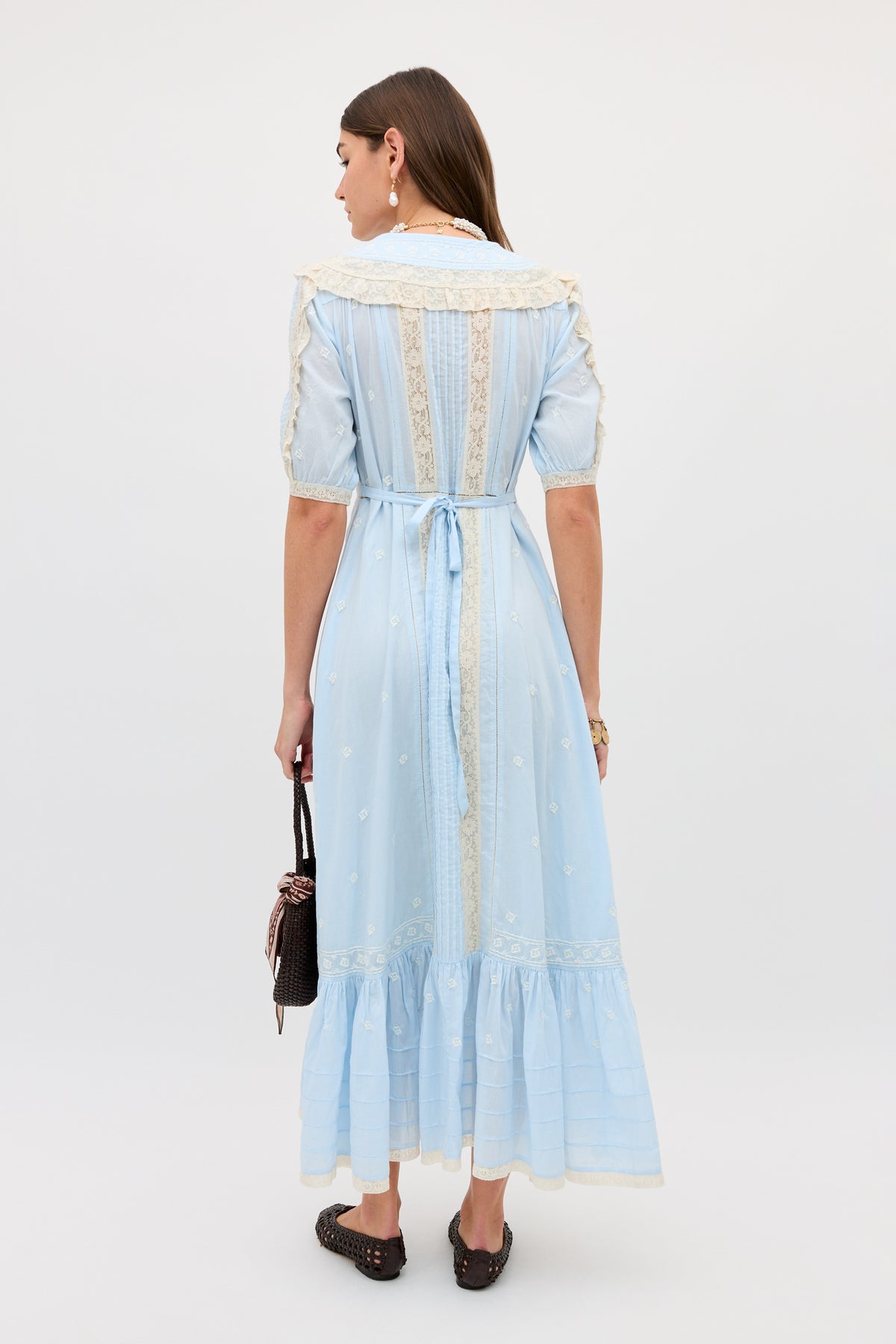 A woman stands facing away in the Edina - Mira Embroidery Powder Blue ankle-length dress with lace trim and waist tie, holding a small black handbag and wearing black slip-on shoes. Her brown hair is down against a plain white background.