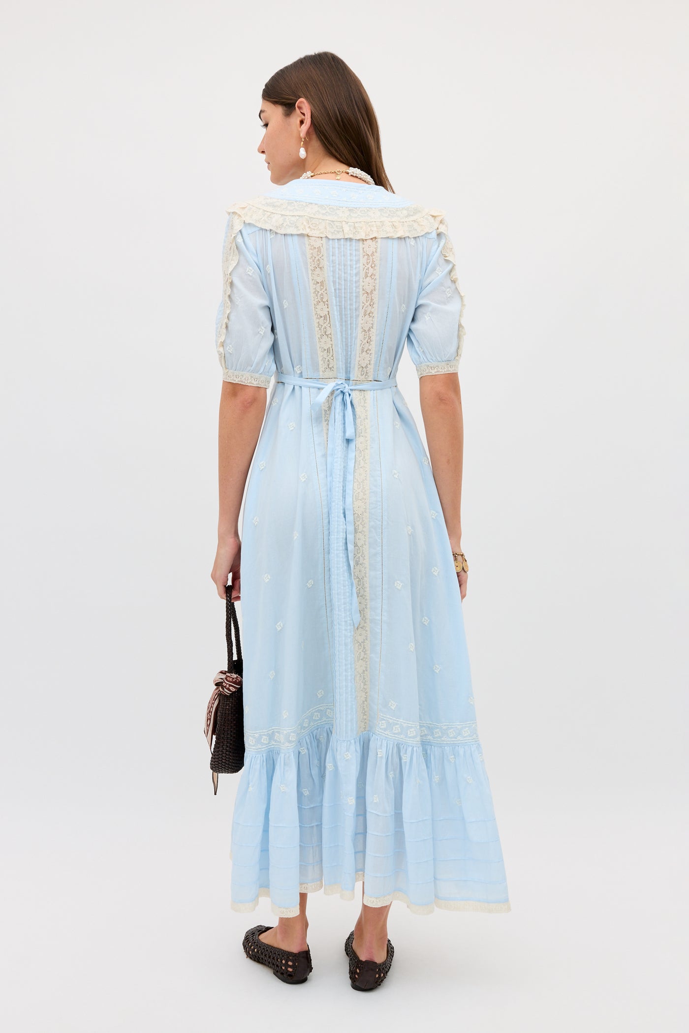 A woman stands facing away in the Edina - Mira Embroidery Powder Blue ankle-length dress with lace trim and waist tie, holding a small black handbag and wearing black slip-on shoes. Her brown hair is down against a plain white background.