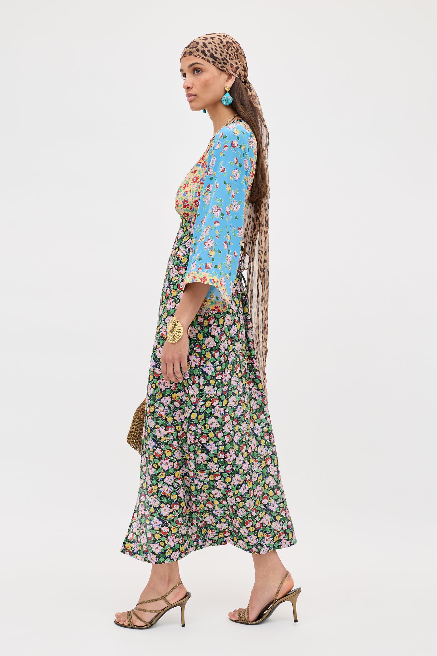 A woman wears the Dahlia - Jeannie Mix Multi, a long dress with blue sleeves and a mixed floral pattern. She pairs it with heeled sandals, a brown patterned headscarf, turquoise earrings, and carries a woven bag against a white background.