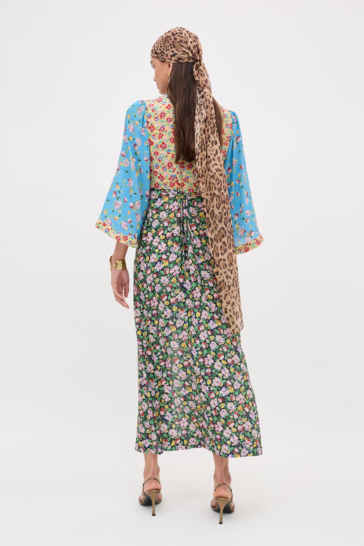 A woman faces away in the Dahlia - Jeannie Mix Multi dress, featuring blue, red, and black floral patchwork. She wears a long leopard print scarf on her head and heeled sandals against a plain white background.