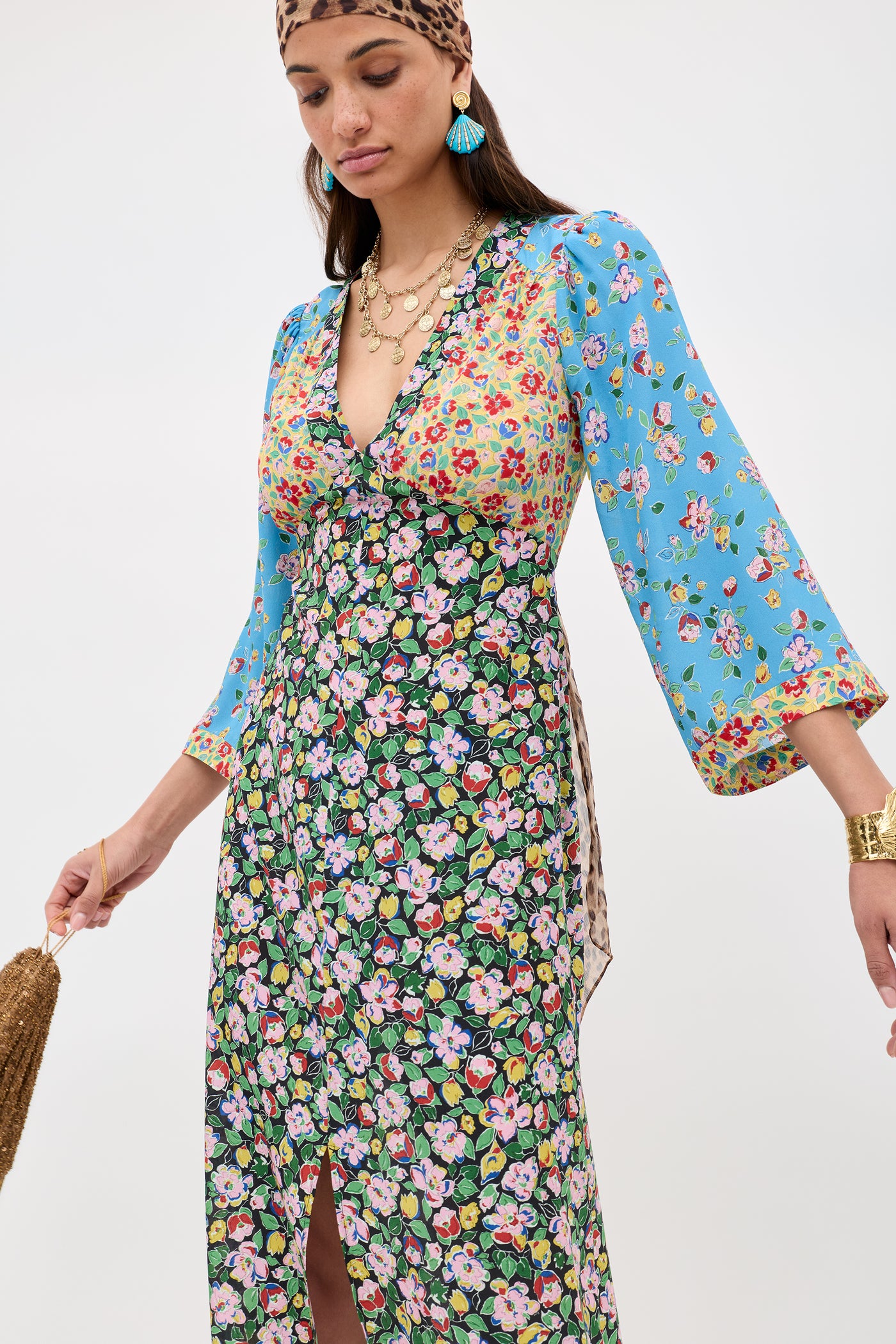 A woman stands against a white background wearing the Dahlia - Jeannie Mix Multi dress, a long mixed floral print with blue floral sleeves and a V-neck, styled with layered necklaces, gold bracelet, teal tassel earrings, and a brown patterned headband.