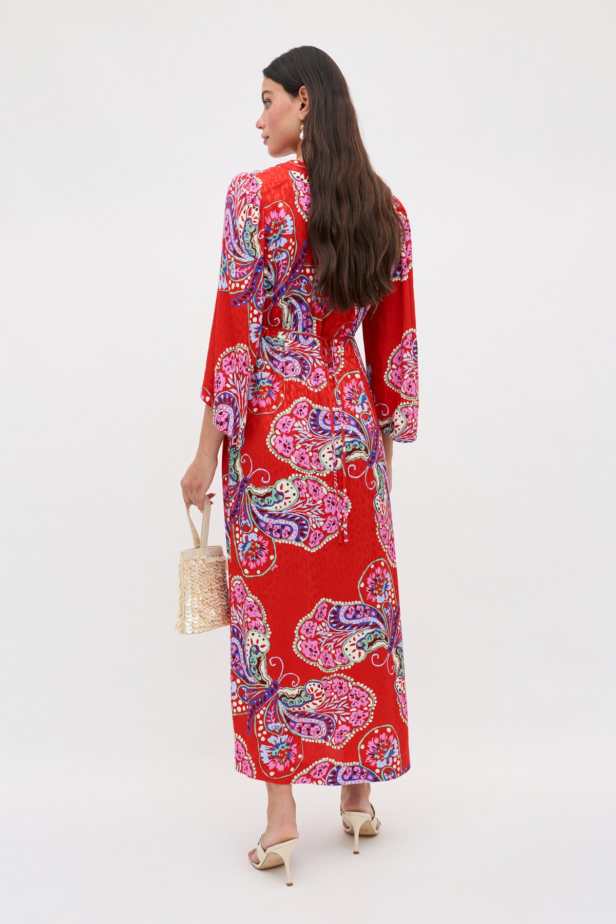 A woman with long brown hair, seen from the back, wears the Dahlia - Butterfly Bloom Jacquard Red dress with cream heeled sandals and holds a straw handbag against a plain light background.