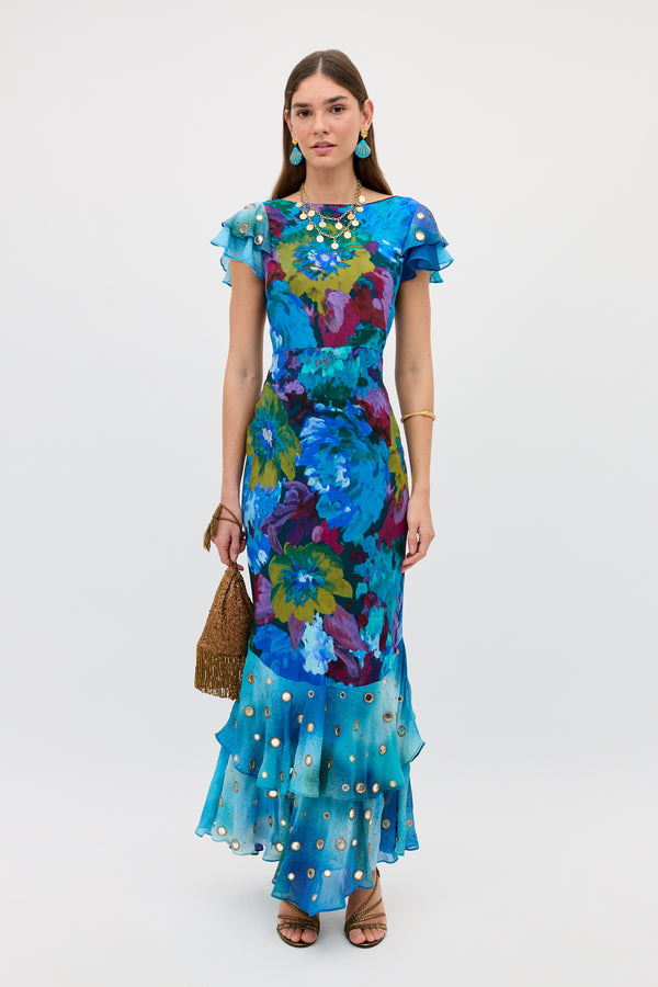 A woman wears the Ordinia - Jessica Bouquet Mix Blue, a fitted floor-length blue floral gown with ruffled short sleeves and hem, gold embellishments, Shisha embroidery, sandals, gold jewelry, and holds a woven handbag against a white background.