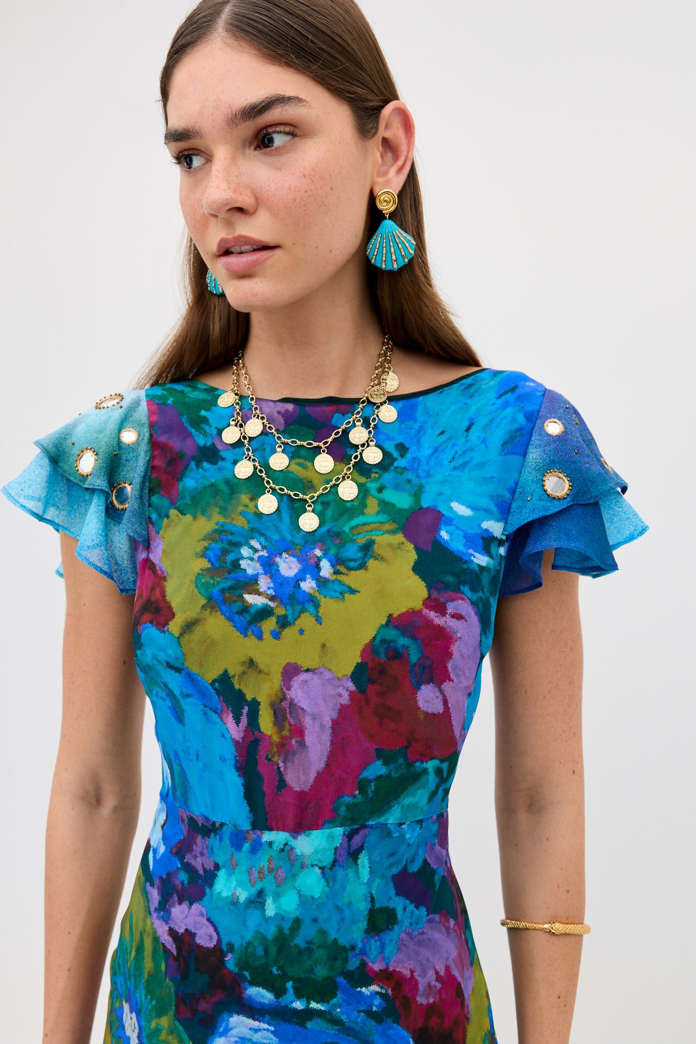 Woman wearing the Ordinia - Jessica Bouquet Mix Blue dress, featuring a vibrant Liberty London floral print and layered ruffle sleeves, styled with gold jewelry and blue tassel earrings as she looks to the side against a plain background—perfect for black-tie weddings.