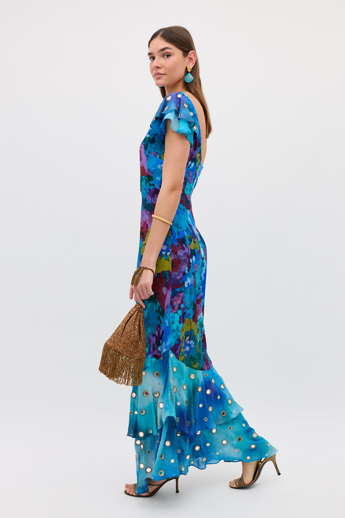 A woman in the Ordinia - Jessica Bouquet Mix Blue ruffled floral maxi dress with sequin accents and Shisha embroidery stands against a plain white background, accessorized with a brown fringed bag, turquoise earrings, and gold heels.