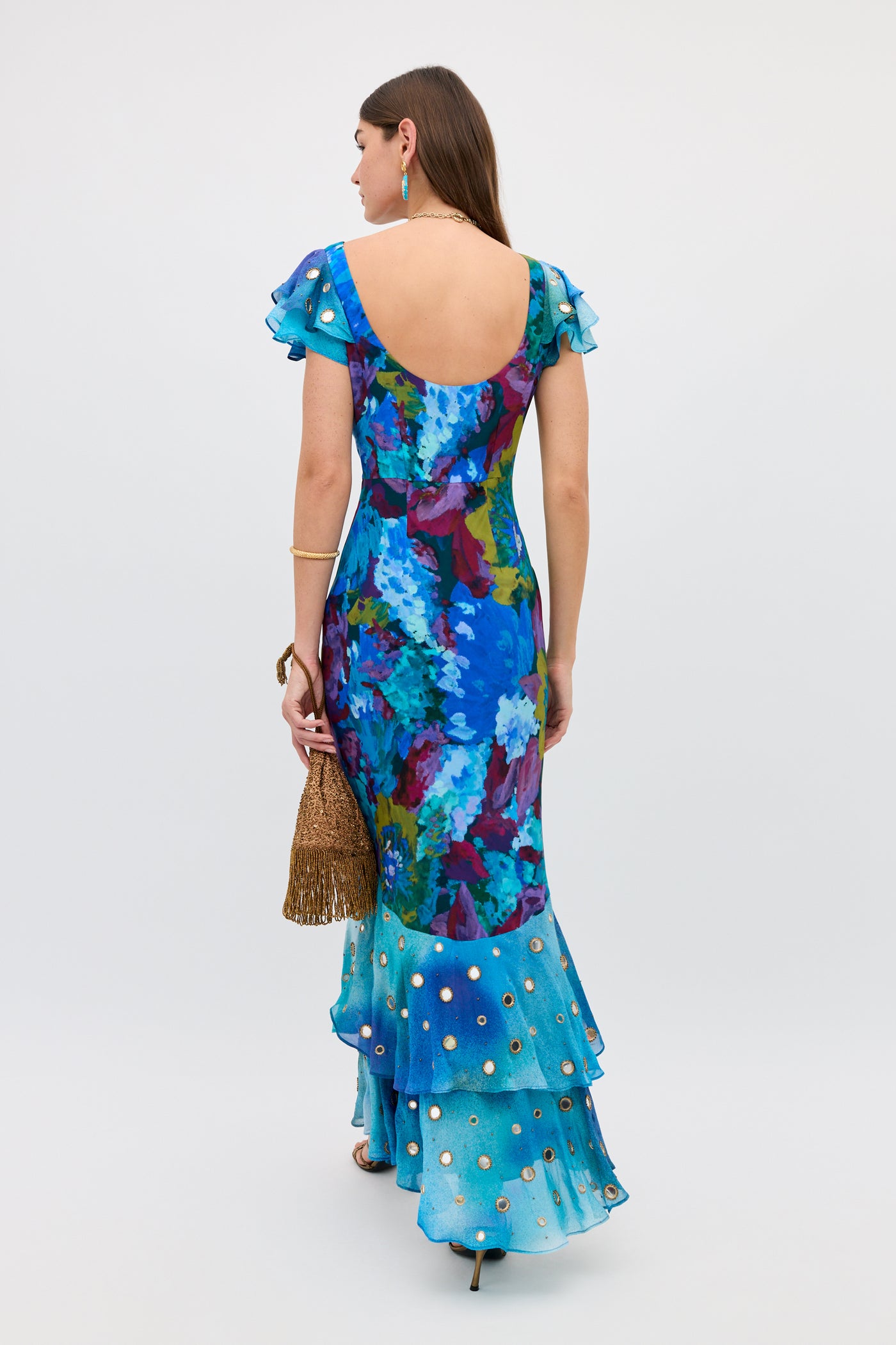 A woman faces away, wearing the Ordinia - Jessica Bouquet Mix Blue gown, a vibrant blue floral dress featuring a ruffled, tiered hem and gold embellishments. She holds a gold tasseled purse. Crafted from a striking Liberty London print, her ensemble is perfect for black-tie weddings. She has long dark hair and accessorizes with earrings and a bracelet.