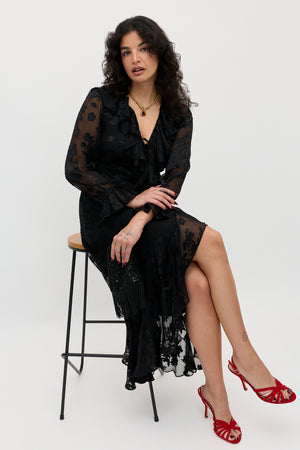 A woman wearing the Gracye – Bouquet Devoré Black, a sheer black dress adorned with floral patterns, sits on a stool with her legs crossed. She accessorizes with red high-heeled sandals and gold jewelry, her wavy dark hair framing a neutral expression against a plain background.