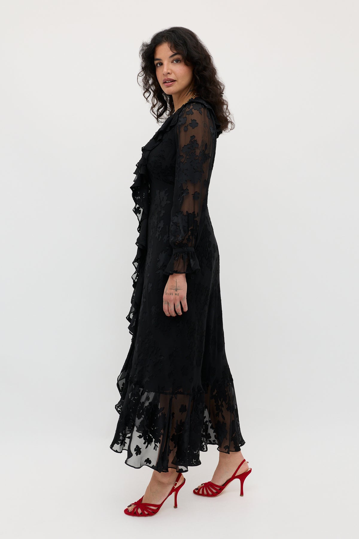 A woman wearing the Gracye - Bouquet Devoré Black, a long, sheer black floral dress with ruffles, stands sideways against a plain white background. She has wavy dark hair and is styled with red high-heeled sandals.