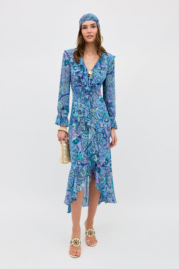 A woman in the Gracye - Paisley Floral Blue long-sleeve ruffled dress with a matching headscarf, white sandals, and a woven handbag stands against a plain light background.