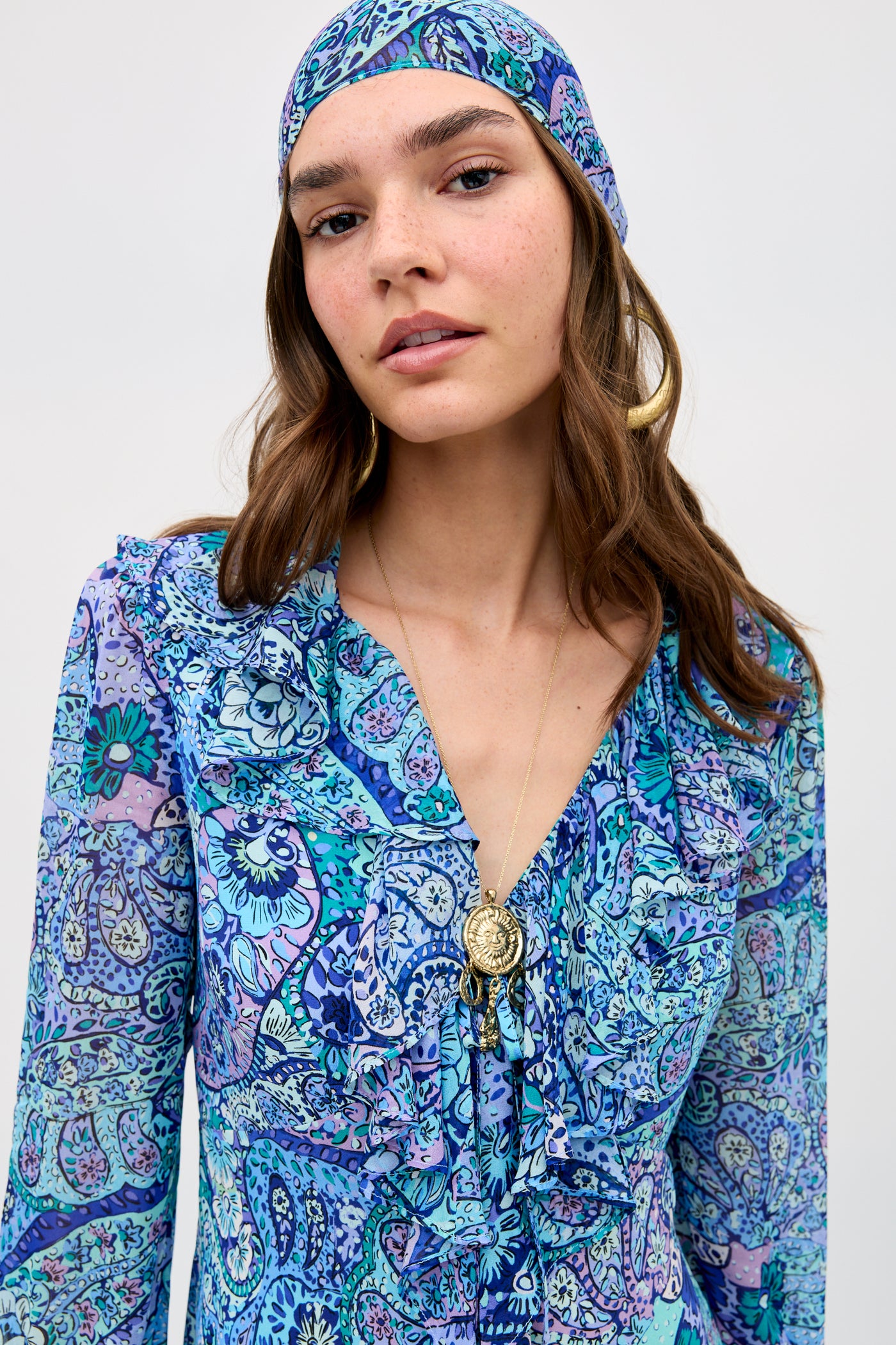 A woman with long brown hair wears the Gracye - Paisley Floral Blue dress and matching headscarf, paired with gold hoop earrings and a pendant necklace, standing against a plain light background.