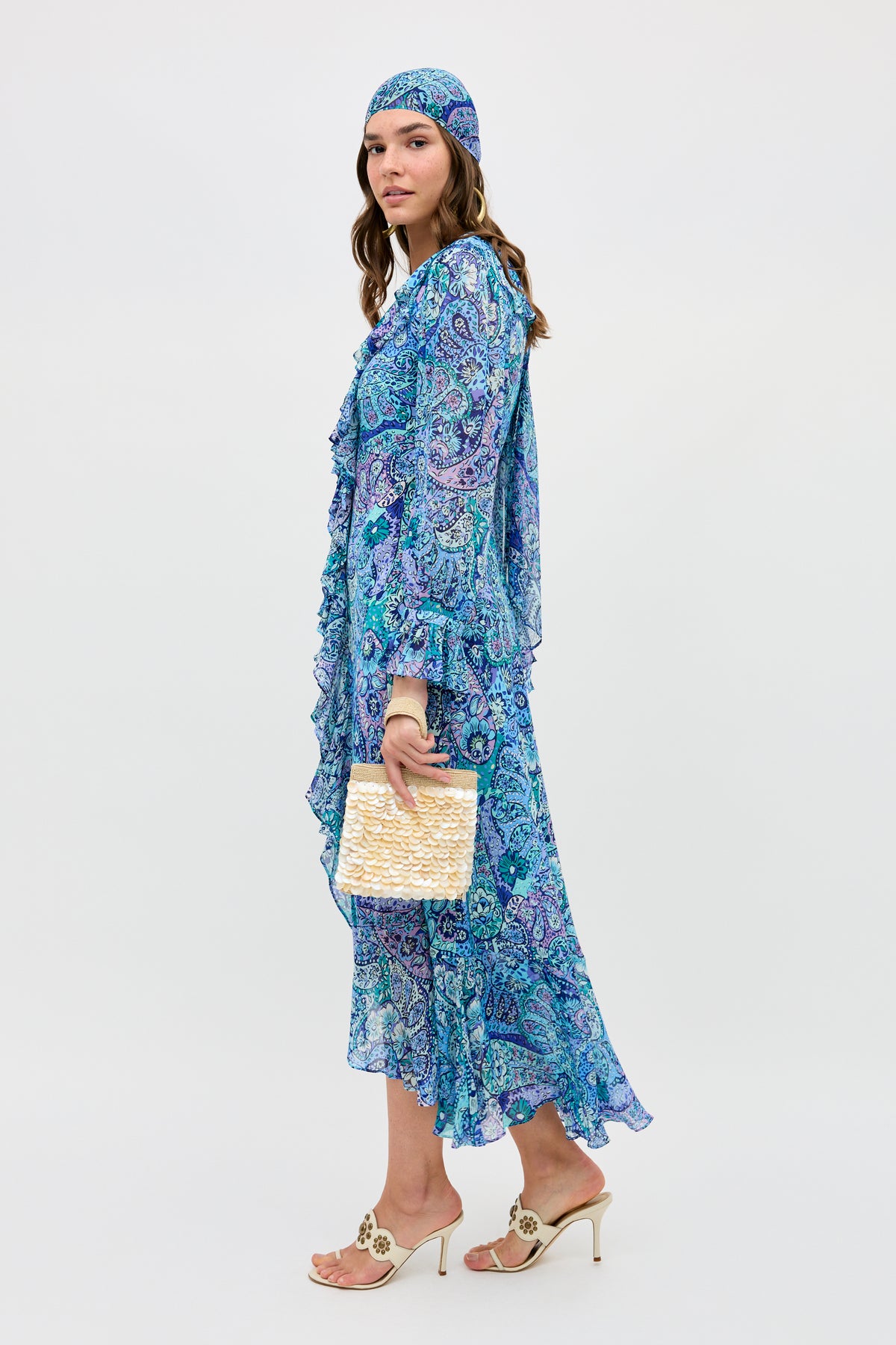 A woman models the Gracye - Paisley Floral Blue dress with a matching headscarf, beige heeled sandals, and a woven beige clutch, standing against a plain white background.