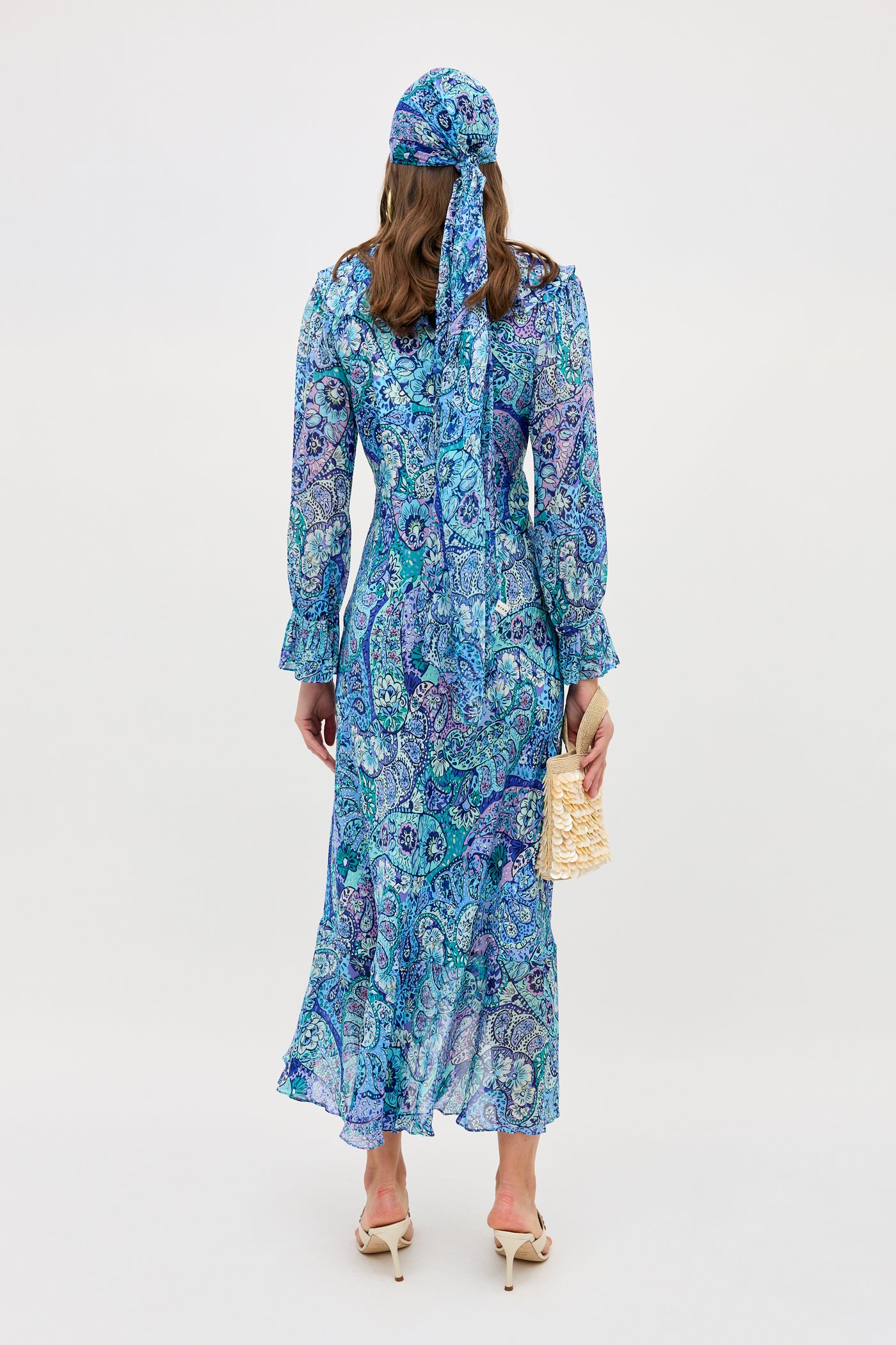 A woman with wavy hair stands facing away against a white background, wearing the Gracye - Paisley Floral Blue dress and headscarf, white heeled shoes, and holding a beige woven handbag.