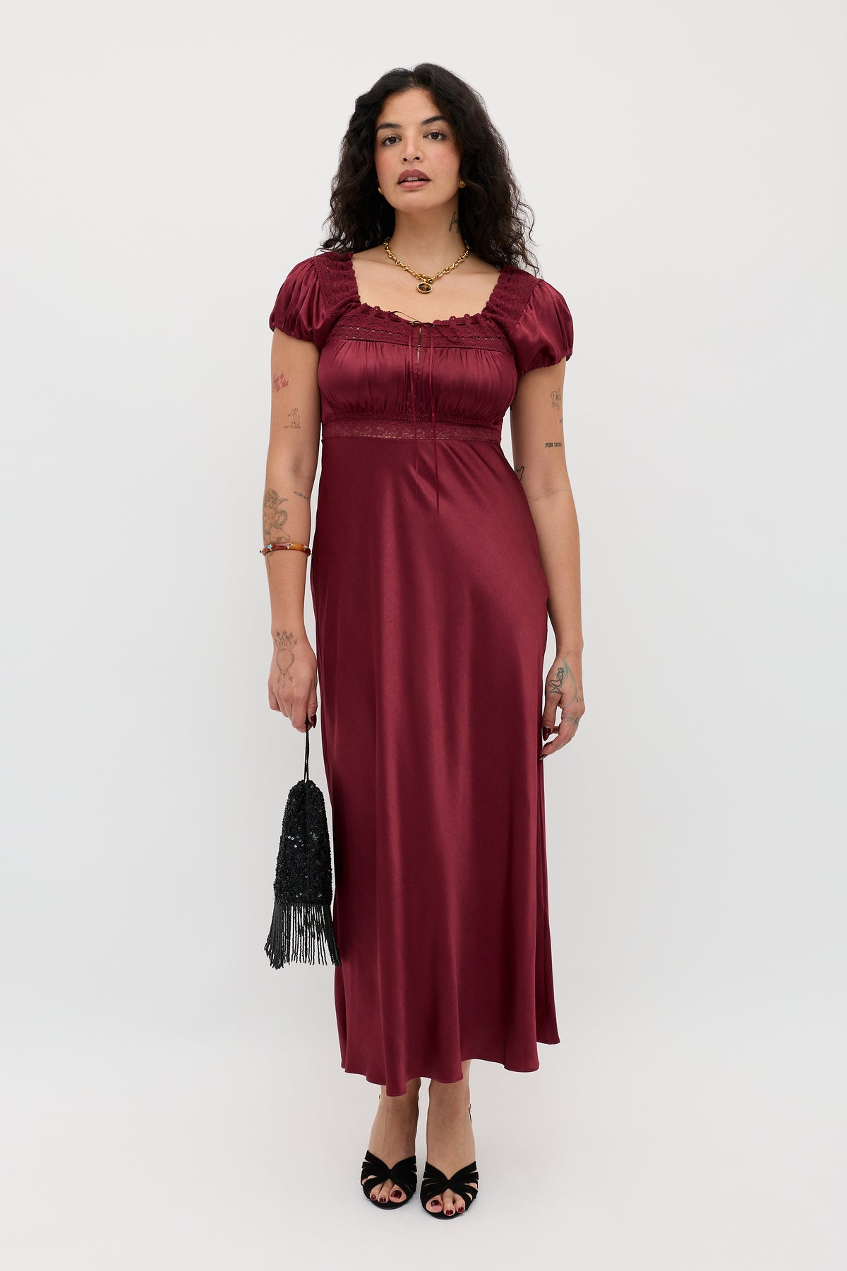 A woman with wavy dark hair wears the Maeva - Maroon shiny short-sleeve maxi dress. She pairs it with a black fringed bag, black open-toe heels, and shows arm tattoos while standing against a plain white background.