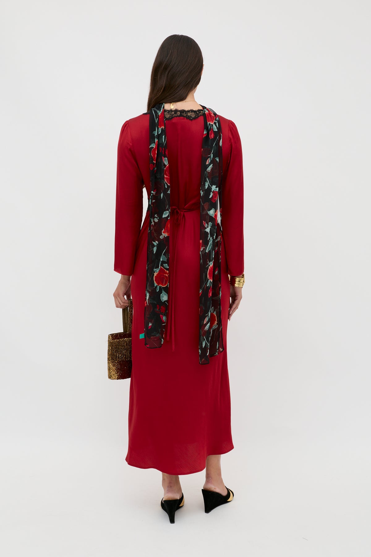 A woman with long brown hair stands facing away, wearing a red dress with a black floral scarf. She carries the Debra - Burgundy textured handbag and completes her look with black heeled mules.
