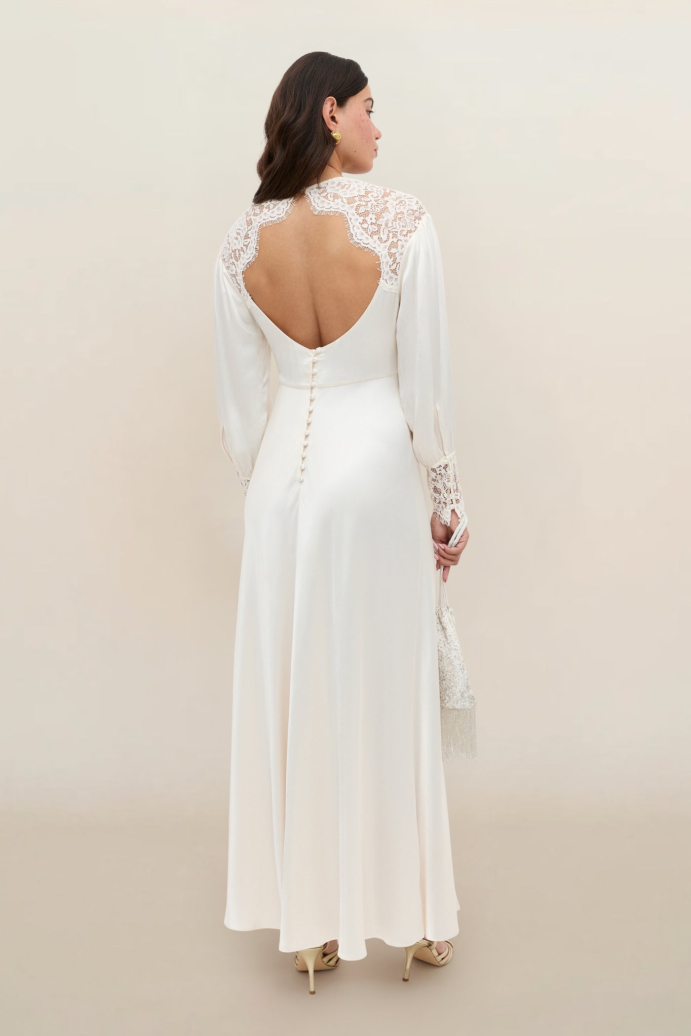 A woman in the Axelle - Pearl gown, an elegant long white dress featuring lace details, an open back, and a row of buttons down the center. She stands facing away, showcasing the stunning back of the dress against a neutral background.