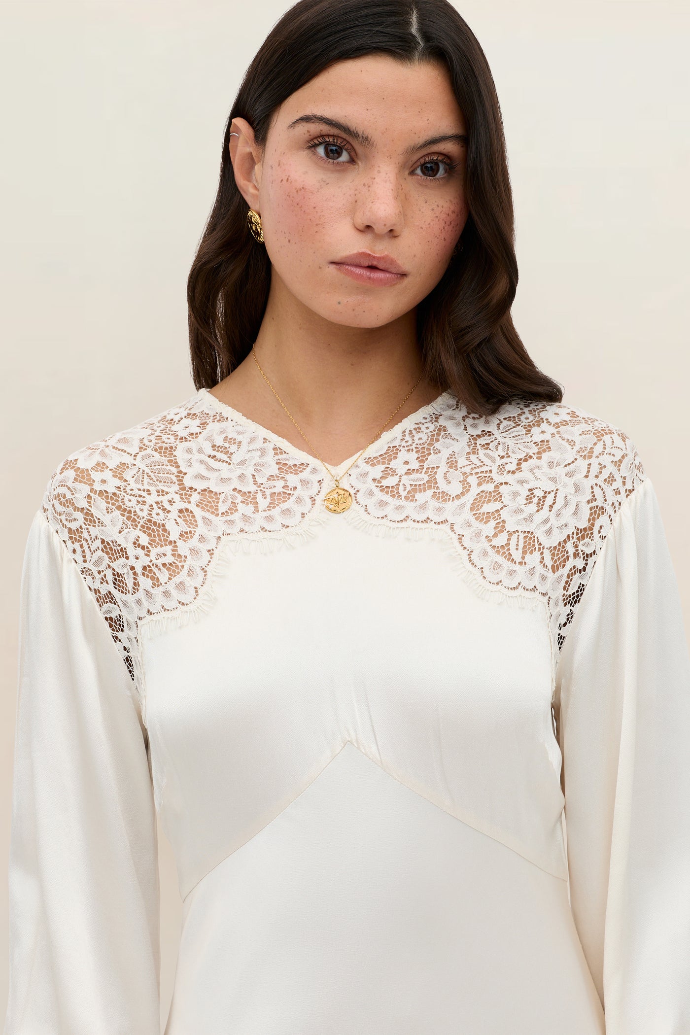 Wearing the Axelle - Pearl ivory dress adorned with lace details on the shoulders and chest, a woman with long brown hair stands against a neutral background, accessorized with gold earrings and a gold necklace.