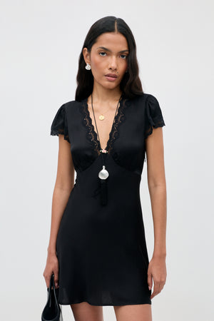 A woman wearing the Cely - Black dress with lace trim, holding a black bag. She has long dark hair, wears earrings and layered necklaces, and stands against a plain light background.