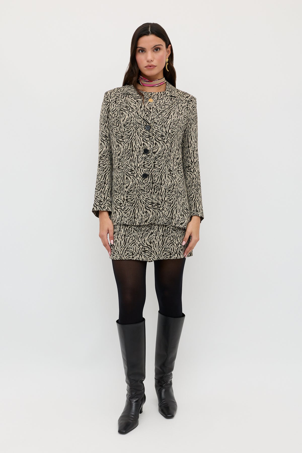 A woman with long dark hair and a neutral expression stands facing forward, wearing the Katrin - Sun Leo Tan patterned blazer and matching skirt, black tights, knee-high black boots, and a red scarf against a plain white background.