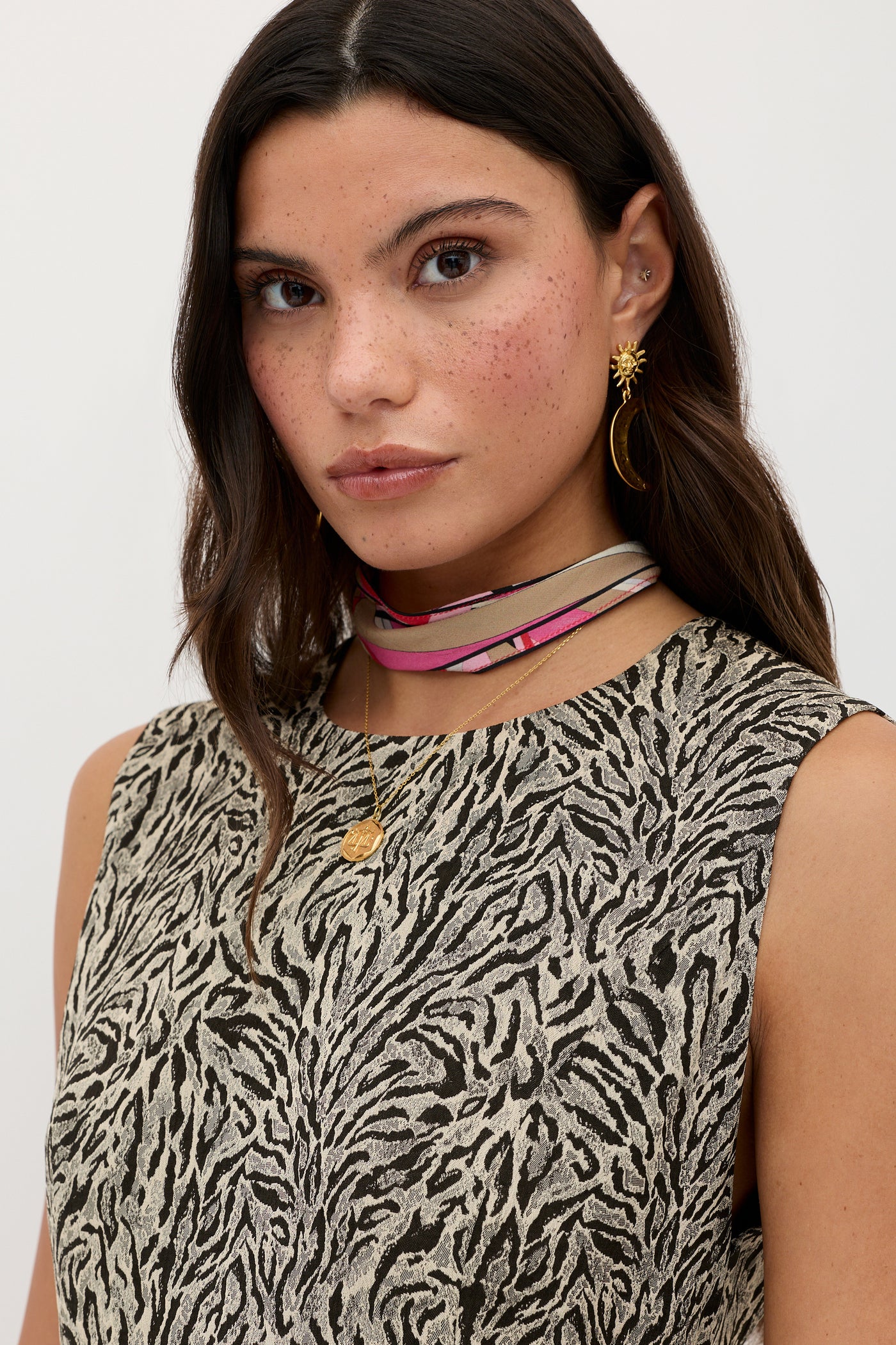 A woman with long brown hair wearing the Katrin - Sun Leo Tan animal-print top, layered choker, gold earrings, and a gold pendant stands against a plain white background.
