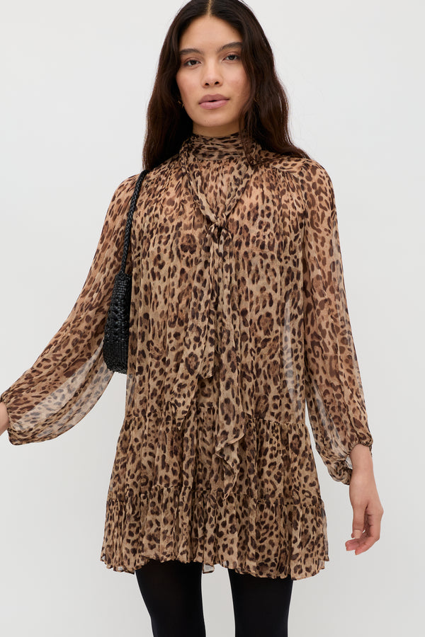 A woman with long dark hair wears the Anaya - Mila Leopard Brown dress, a long-sleeved, above-the-knee leopard print style with a ruffled hem and tied high neckline. She carries a black shoulder bag and stands against a white background.