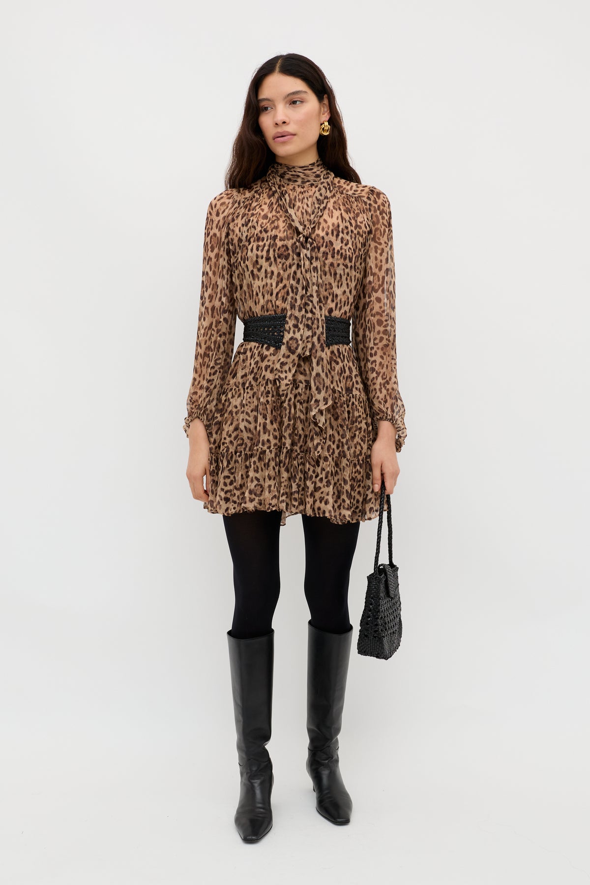 A woman poses against a white background, wearing the long-sleeved, high-neck Anaya - Mila Leopard Brown dress with a black belt, tights, knee-high boots, a black handbag, gold earrings, and long wavy hair for a chic look.