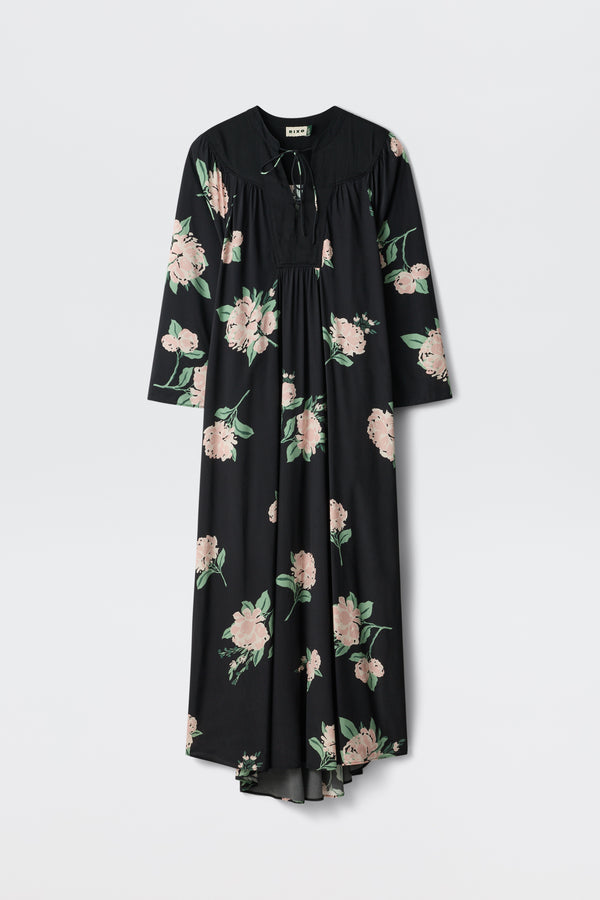 The Lucilla - Peonies Scatter Black, a long-sleeved black maxi dress featuring a pink and green floral pattern, is displayed against a plain white background.