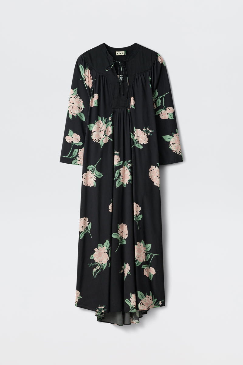 The Lucilla - Peonies Scatter Black, a long-sleeved black maxi dress featuring a pink and green floral pattern, is displayed against a plain white background.