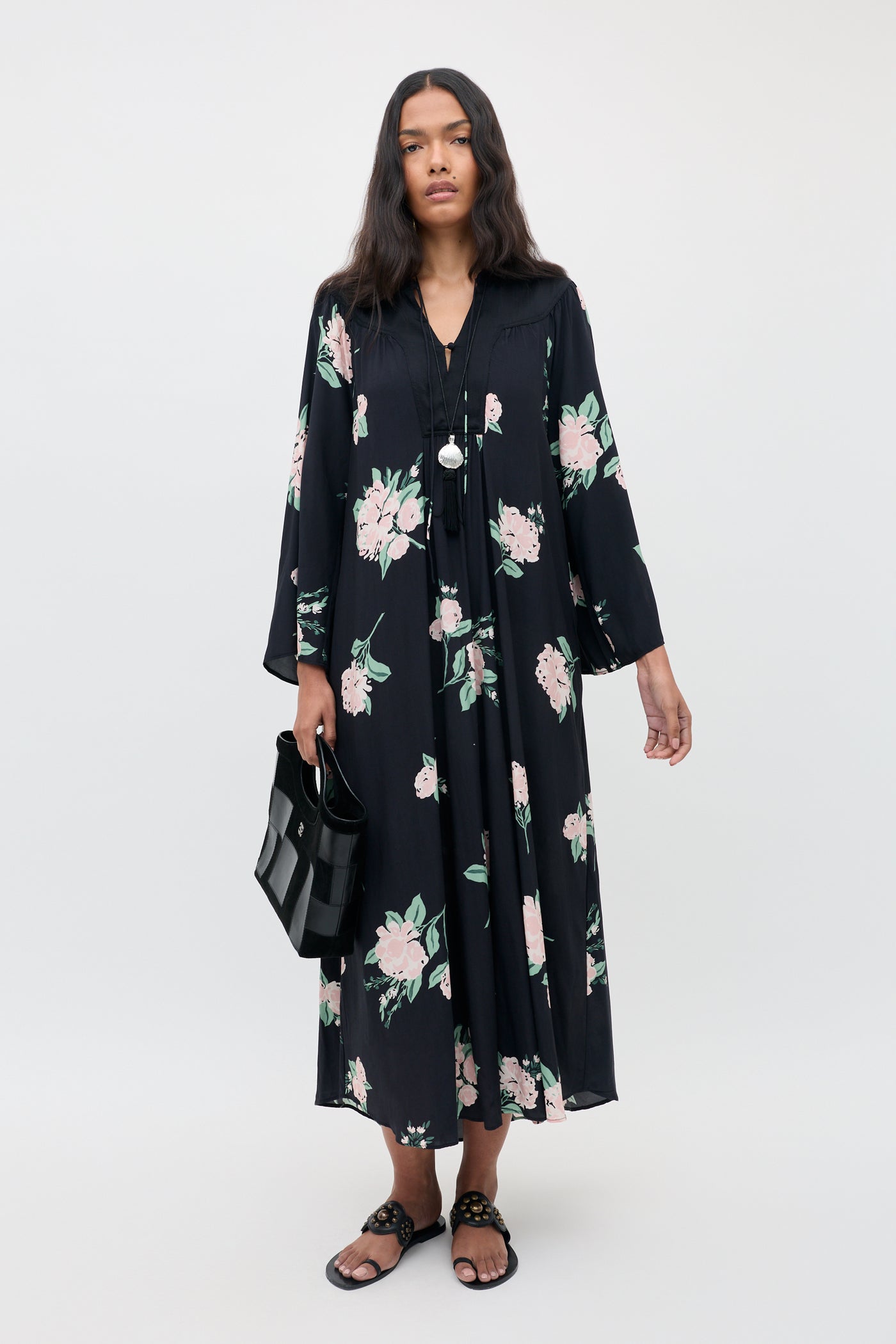 A woman wearing the Lucilla - Peonies Scatter Black dress, featuring pink and green floral patterns, paired with black sandals and a black handbag, stands against a plain light background.