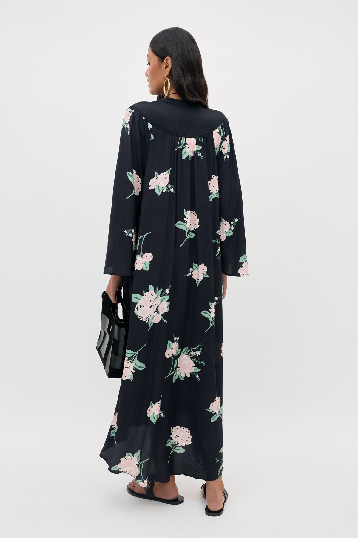 A woman with long dark hair wears the Lucilla - Peonies Scatter Black maxi dress, featuring pink floral patterns and long sleeves, holds a black handbag, and stands against a plain light background.