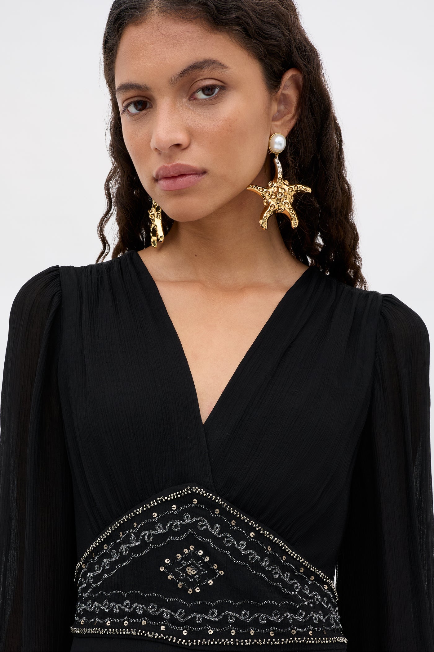 A woman with wavy brown hair wears the Kaesha - Diana Embellishment Black dress, featuring a V-neck and intricate waist beadwork. She accessorizes with gold starfish and pearl earrings, posing neutrally against a light background.