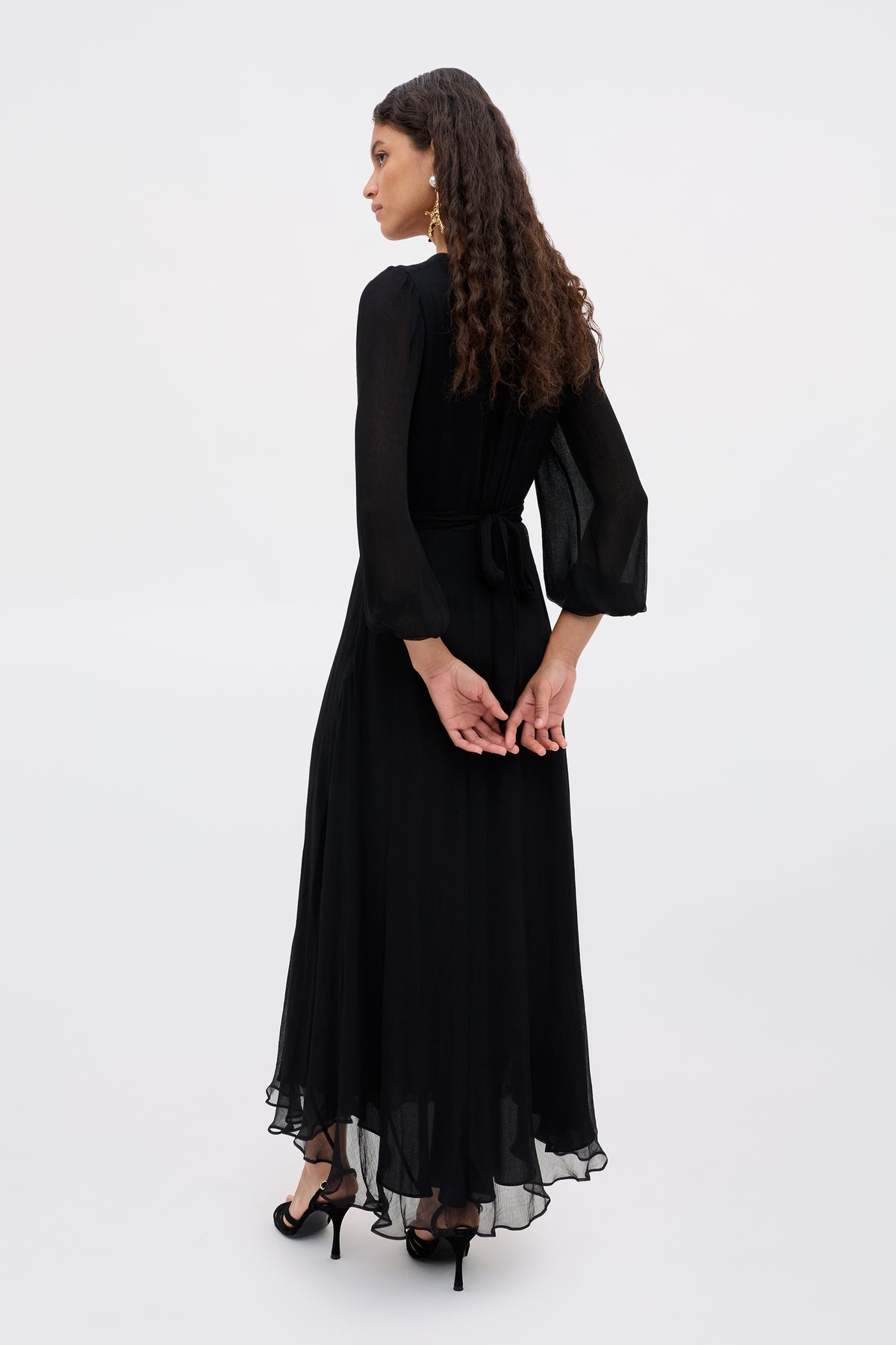 A woman with long curly hair wears the Kaesha - Diana Embellishment Black dress and black heels, standing against a light background, turned slightly away with her hands clasped behind her back.