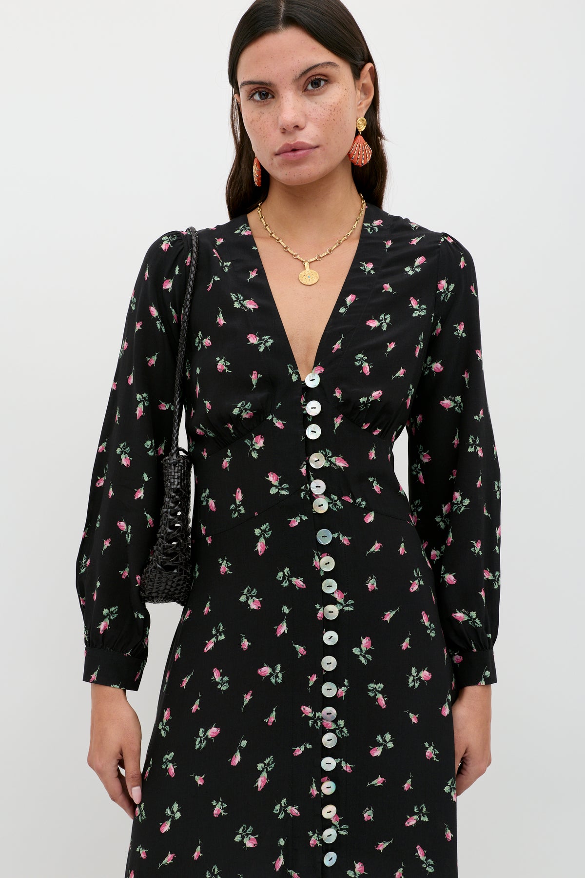 A woman wearing the Avalyn - Sienna Bud Black dress, a black long sleeve midi with a pink floral pattern and vintage cuffs, styled with gold jewelry and a black textured bag, stands against a plain white background.