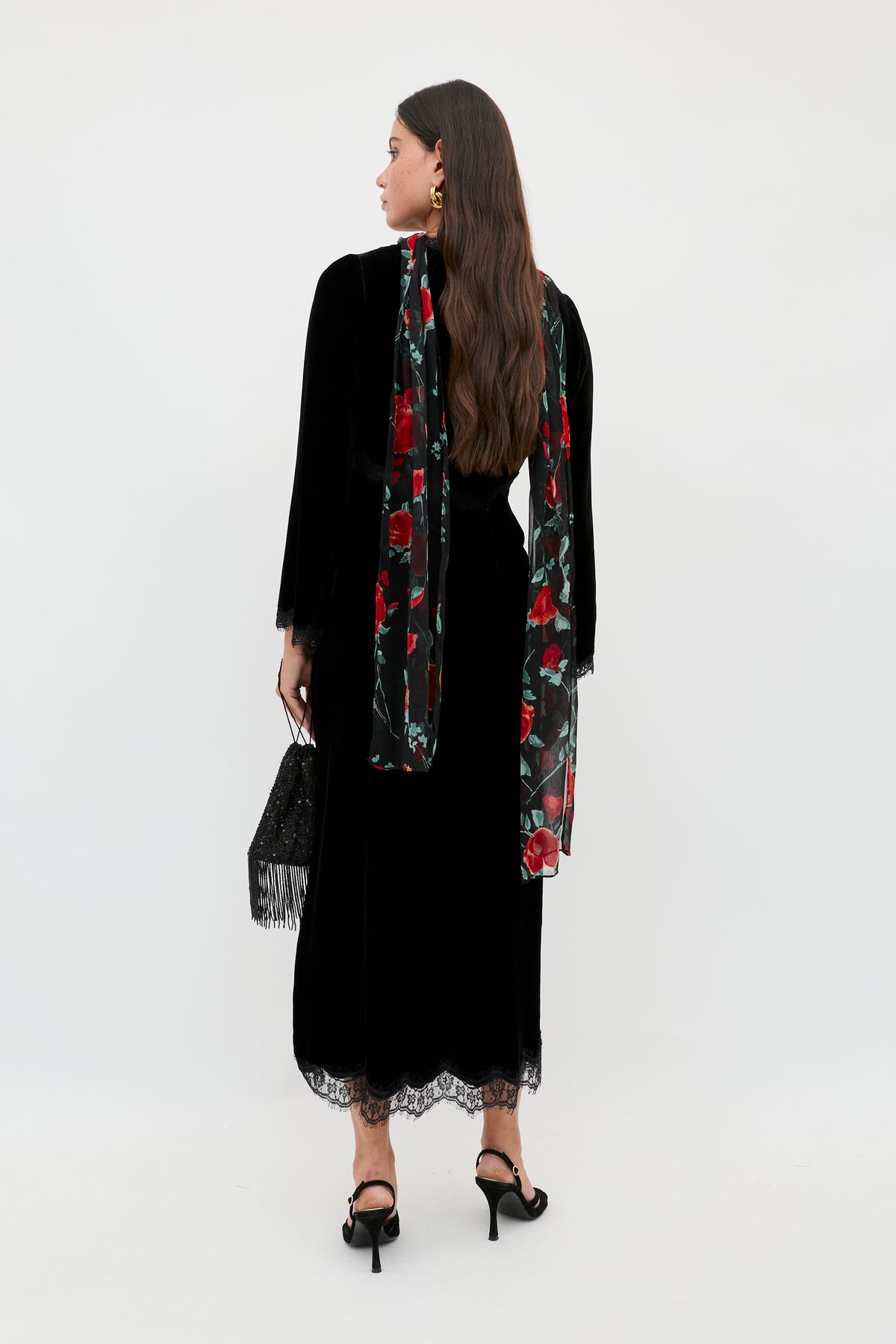 A woman stands against a plain background wearing Alessi - Black, a long black dress with lace detailing, styled with black high-heeled sandals, a red rose floral scarf, and a black handbag. Her long brown hair completes the boho-glamour look.
