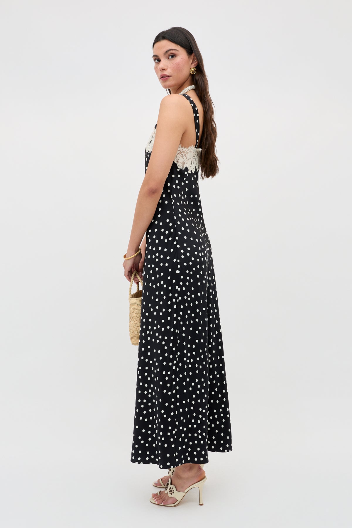 A woman stands sideways wearing the Rosamarie - Scattered Dot Black dress with white lace trim, holding a small woven bag and wearing white heeled sandals. She has long brown hair against a plain, light-colored background.
