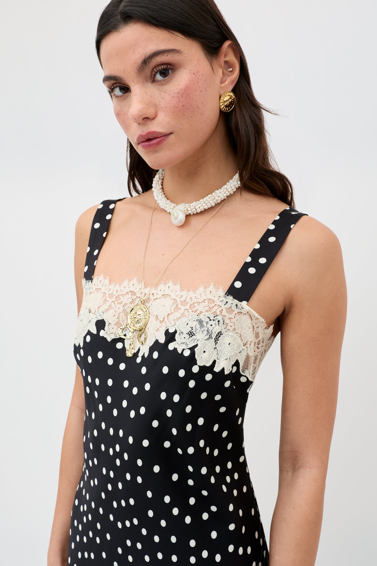 Wearing the Rosamarie - Scattered Dot Black dress with white polka dots and lace trim, a woman accessorized with layered necklaces and gold earrings stands against a plain light background, gazing slightly to the side.