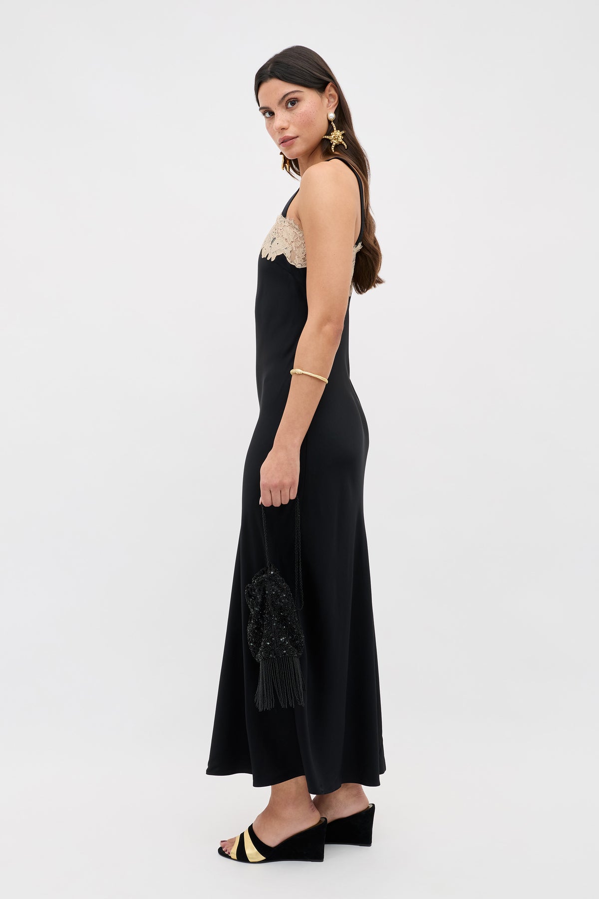 A woman stands sideways in a Rosamarie - Black sleeveless dress with a beige lace top, holding a black beaded purse and wearing black and gold open-toe heels, gold earrings, and a bracelet against a plain white background.