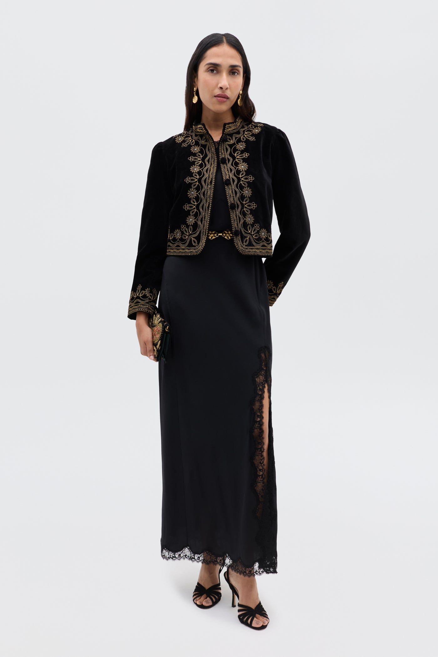 Wearing the Mirena - Black maxi dress with a high slit and lace trim, paired with a black velvet jacket with gold embroidery, black heels, and gold earrings, a woman stands against a plain light background.