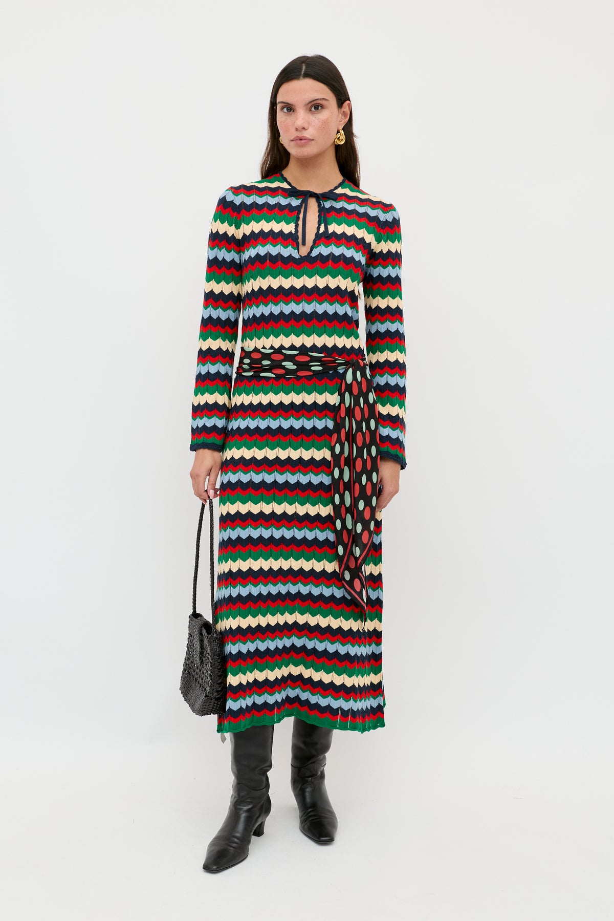 A woman is wearing the Lucila - Retro Chevron Blue long-sleeved dress, featuring a multicolored wavy chevron pattern and a keyhole neckline, styled with a patterned belt, black boots, and a black textured handbag, standing against a plain white background.