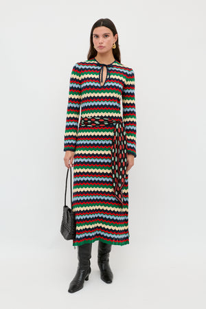 A woman is wearing the Lucila - Retro Chevron Blue long-sleeved dress, featuring a multicolored wavy chevron pattern and a keyhole neckline, styled with a patterned belt, black boots, and a black textured handbag, standing against a plain white background.